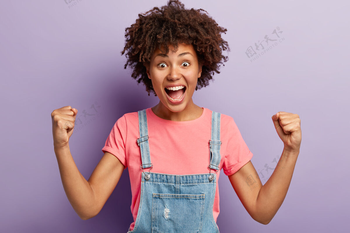 庆祝情绪化的黑皮肤女孩为最喜爱的足球队呐喊 举起紧握的拳头 成为真正的球迷或支持者 穿着休闲服 对着紫色的墙壁摆姿势人民和胜利看满足胜利