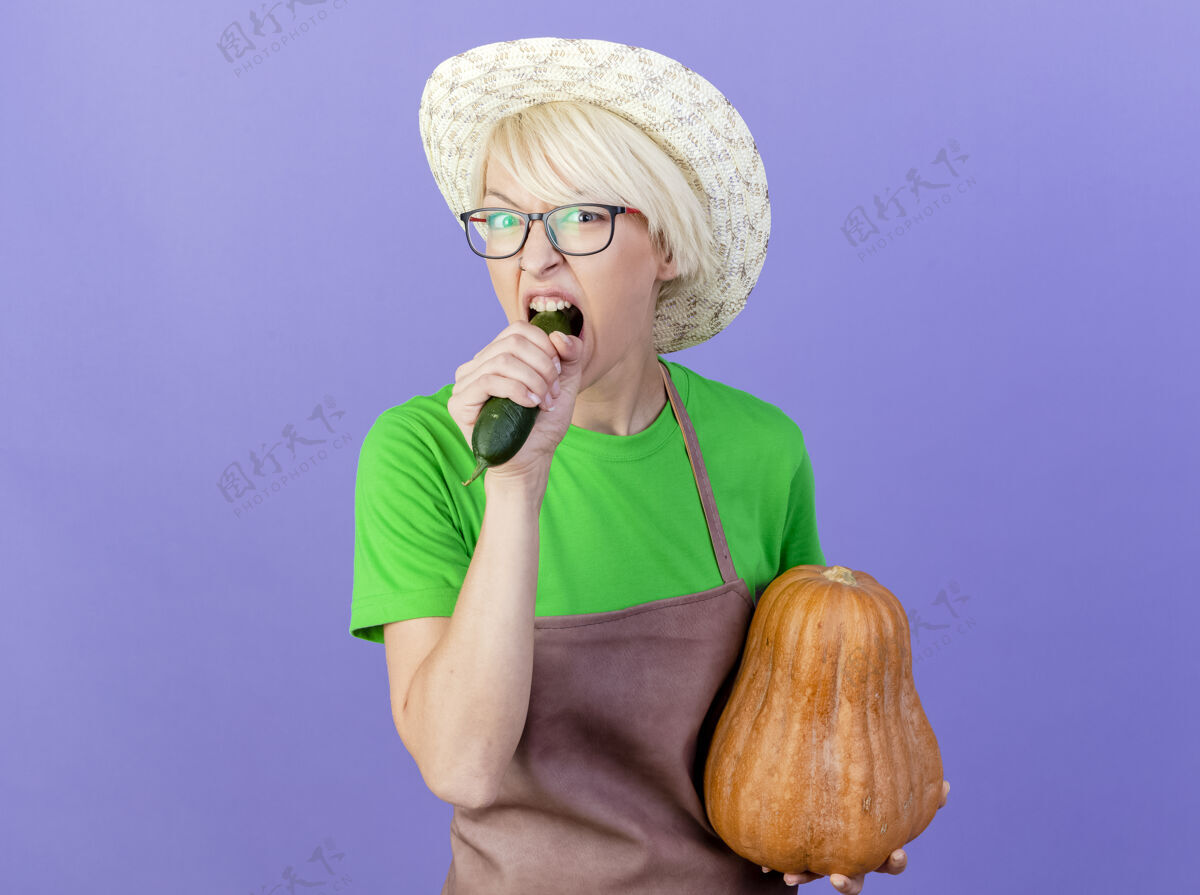 南瓜围裙里留着短发 戴着帽子 手里拿着南瓜咬黄瓜的年轻园丁女人站在蓝色的背景上头发咬女人