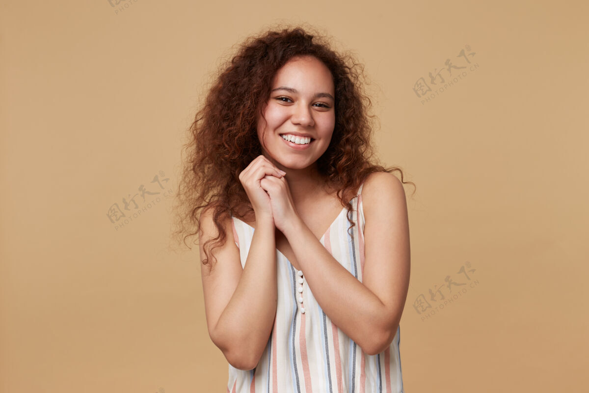 欧式快乐的年轻棕色眼睛卷曲的黑发女人 随意的发型 双手合十 面带迷人的微笑 孤立的米色穿女士休闲