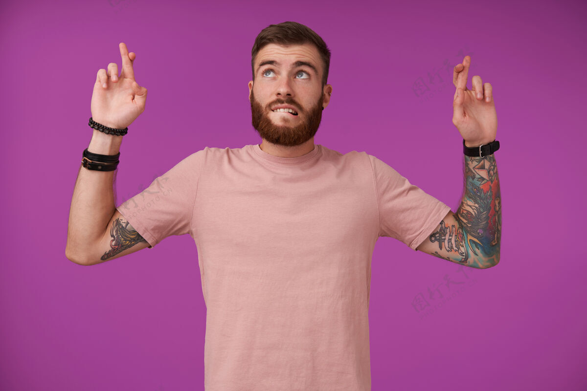 纹身一幅年轻英俊 留着胡须 留着文身 留着短发的黑发男性的肖像 站在紫色的地面上 向上看 咬着臀部 交叉着手指 希望更好无胡子摆姿势情感