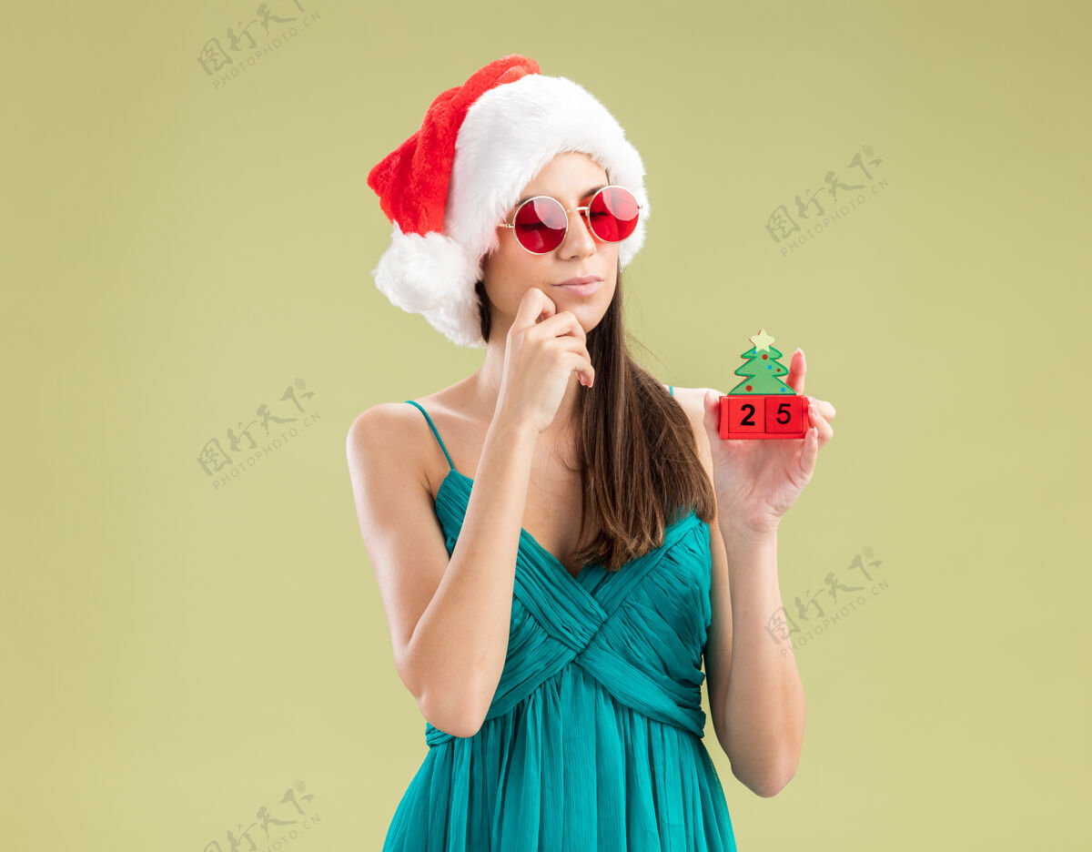 圣诞快乐体贴的年轻白人女孩戴着太阳眼镜 戴着圣诞帽 手放在下巴上 手里拿着圣诞树的装饰品橄榄圣诞老人举行