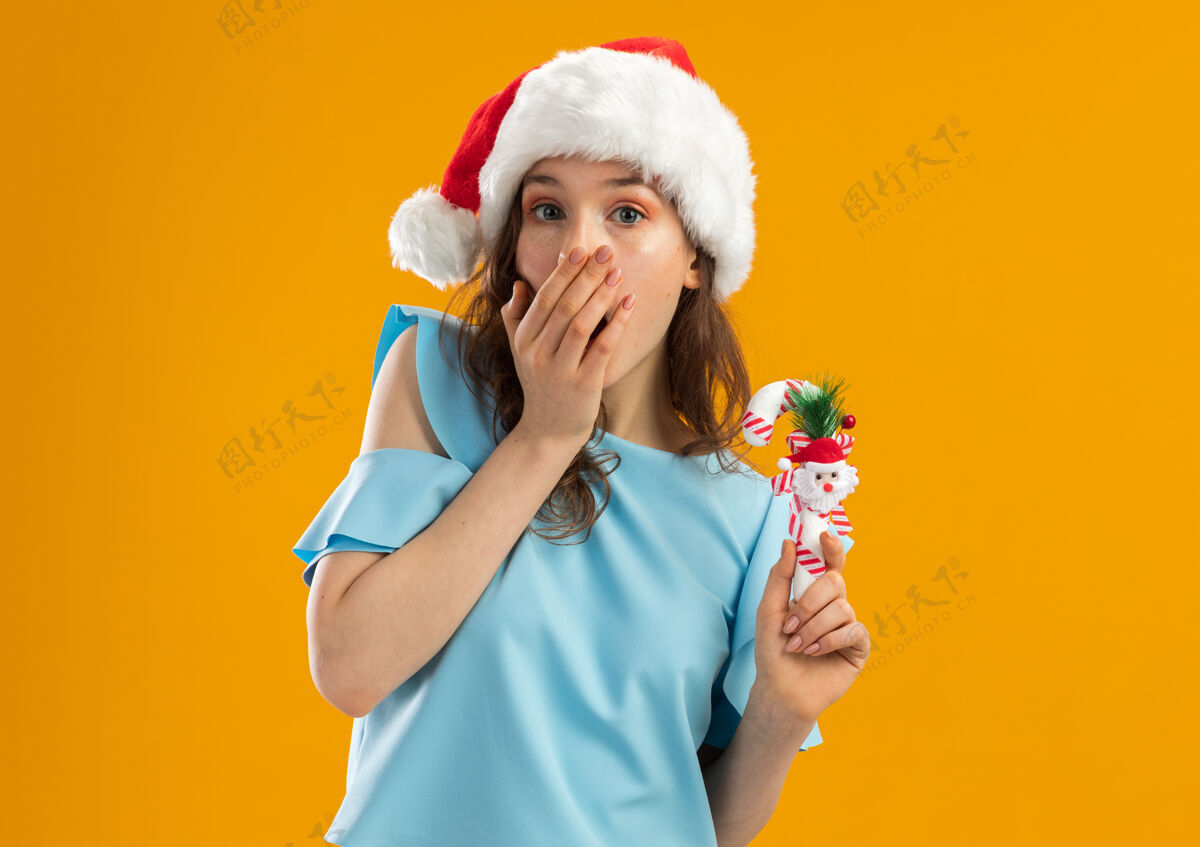 橙色身穿蓝色上衣 戴圣诞帽的年轻女子手持圣诞糖果手杖 用手捂着嘴 看上去很震惊糖果抱着新的