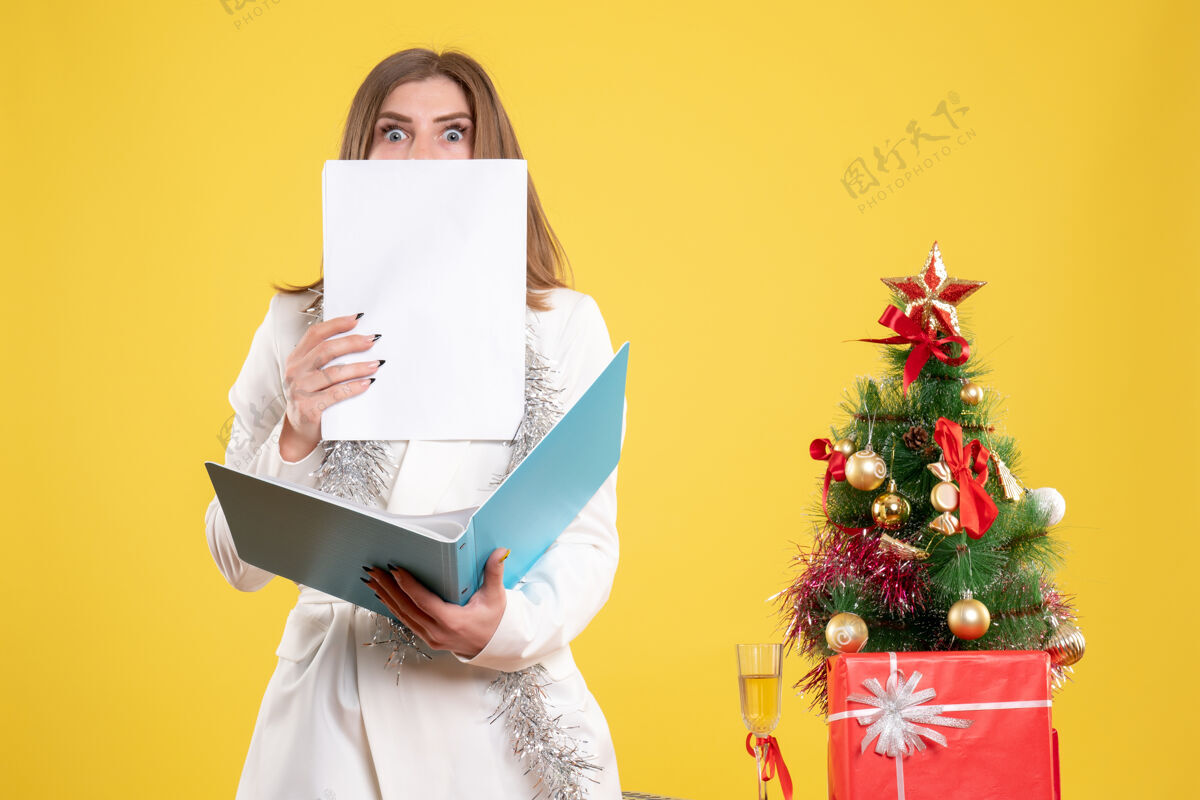背景正面图女医生站着拿着黄色背景上的文件 还有圣诞树和礼品盒持有前面成人