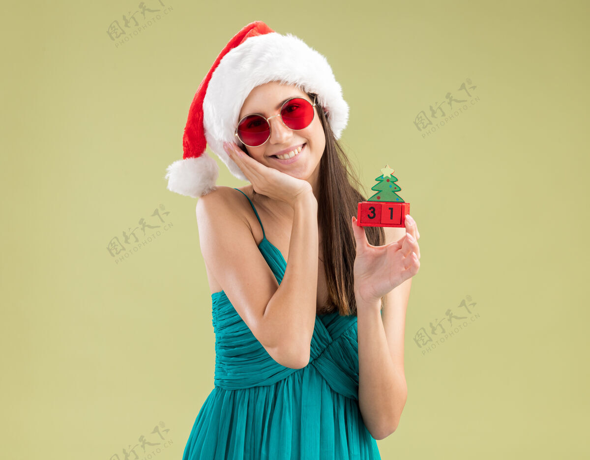 圣诞老人戴着太阳眼镜 戴着圣诞帽的年轻白种女孩高兴地把手放在脸上 手里拿着圣诞树饰物脸眼镜绿色