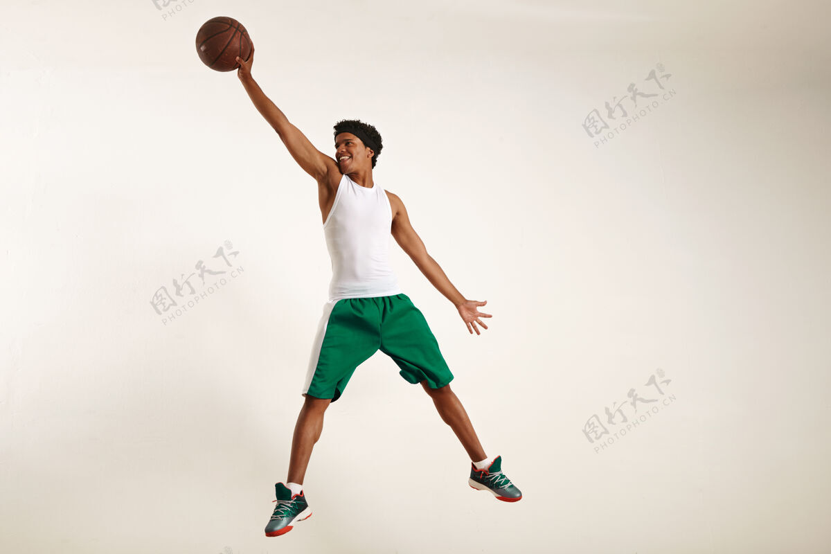 触摸一个穿着白衬衫和绿短裤的快乐的年轻黑人运动员的动作照片跳高抓住一个老式篮球在白色高技术抓