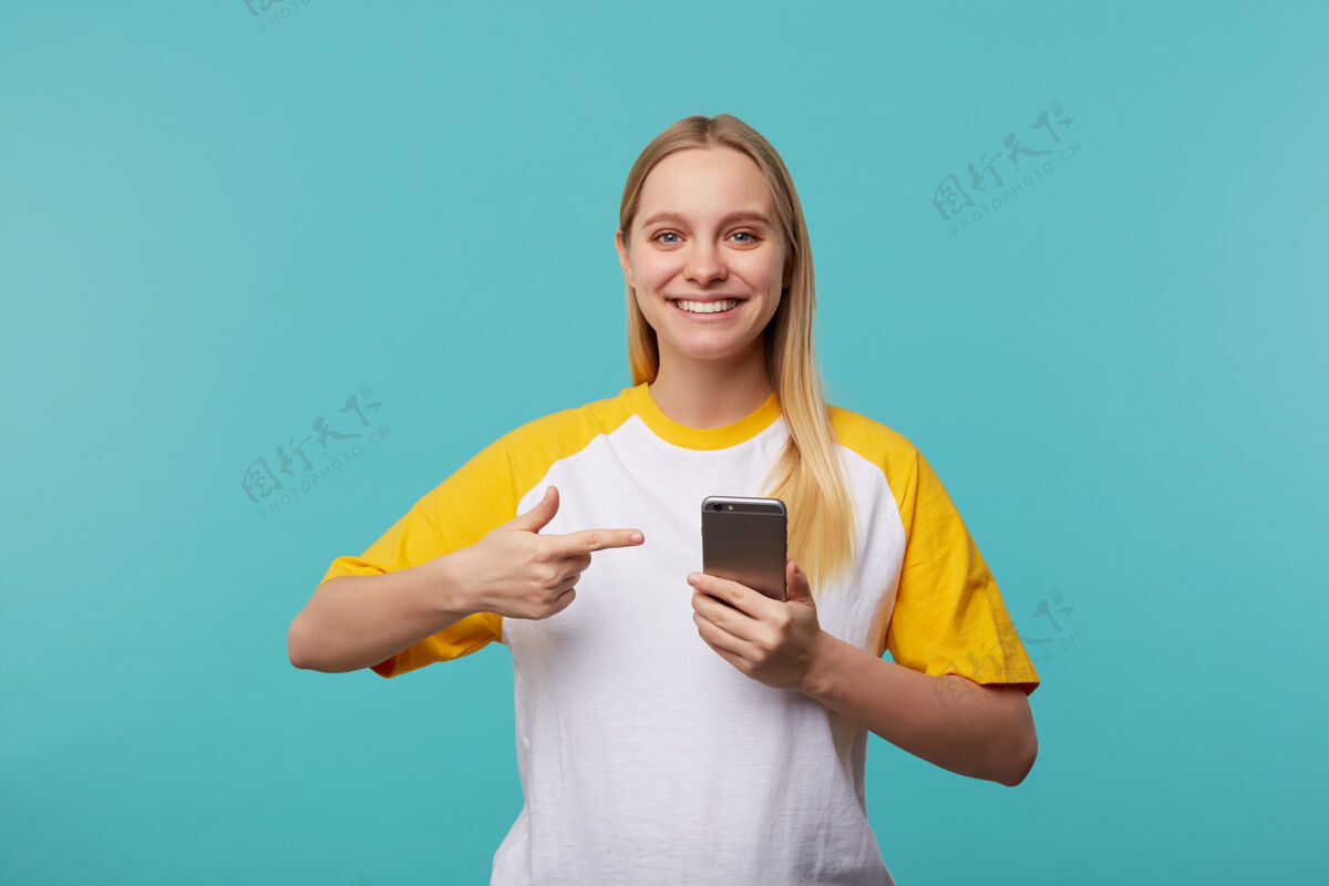 穿着室内写真 年轻迷人的白头女指着手机 开心地笑着 站在蓝色的休闲服上站立手机黄色