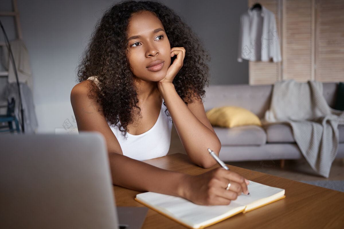 非洲职业 工作 技术和联系体贴 沉思 皮肤黝黑 头发浓密的年轻女子用笔记本电脑做远程工作 在抄写本上写下 试图回忆一些事情成人笔记本女人
