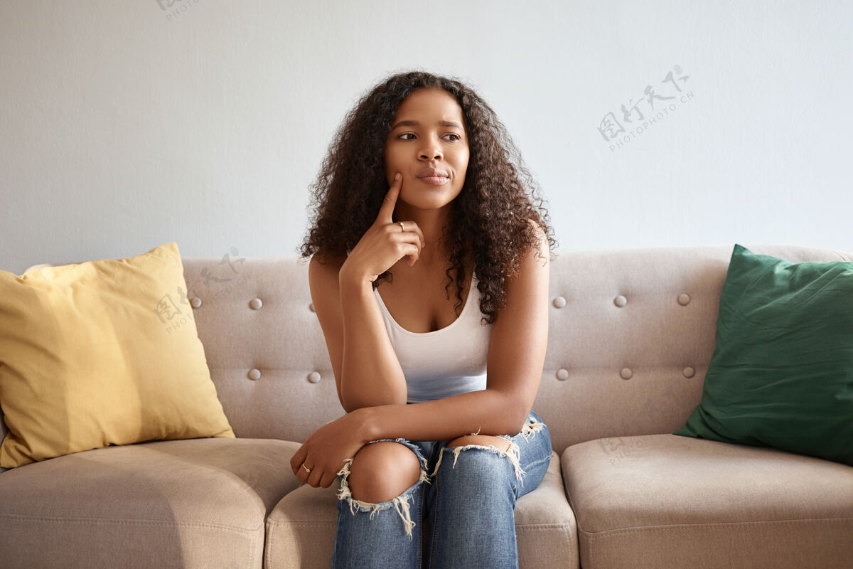 头发室内形象：非洲模样的时尚可爱女孩 孤零零地坐在灰色沙发上 黄绿色的靠垫上 神情可疑而体贴 抚摸着下巴 说：嗯 让我想想年轻思考女人