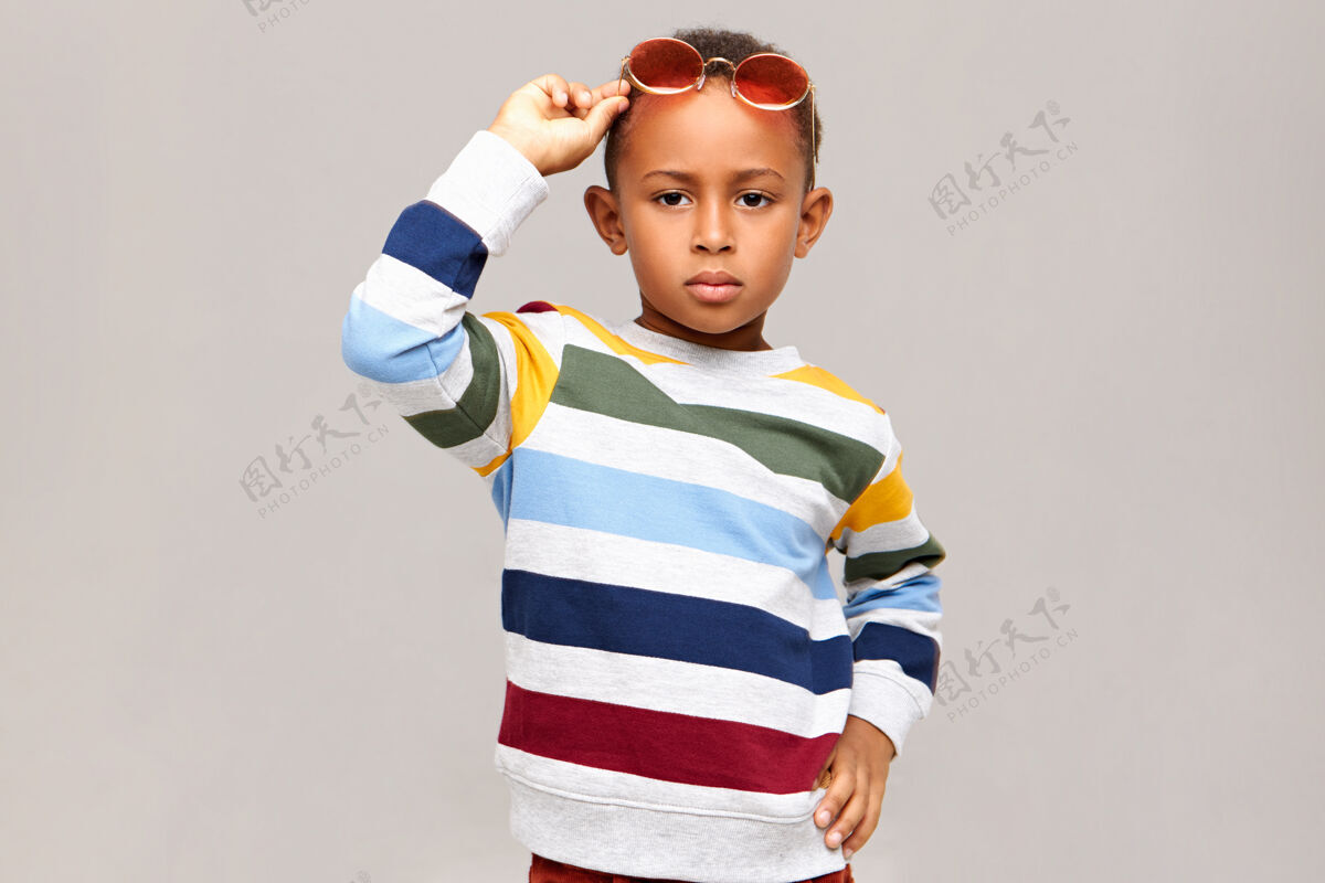小儿童时尚 风格 童装和配饰概念严肃自信的非洲裔美国男孩穿着条纹套头衫 头上戴着粉色的墨镜 在空白的墙壁上做模特年轻小孩子