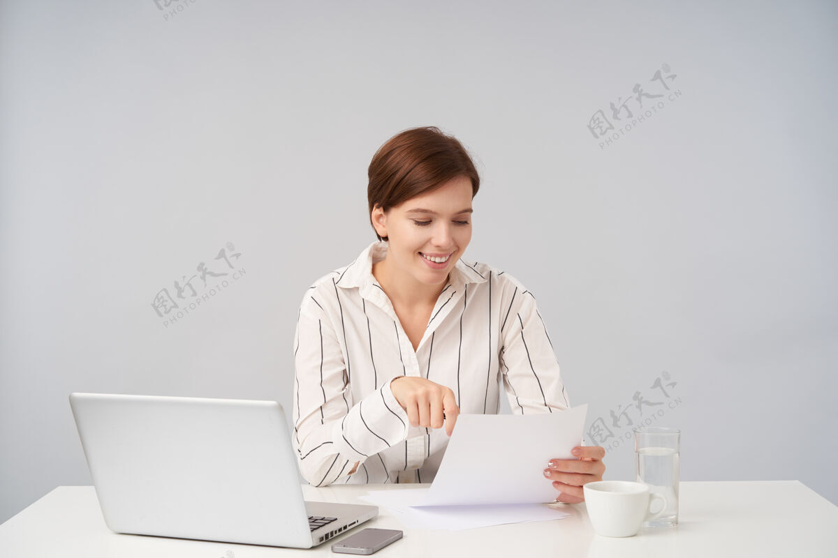 办公室快乐的年轻漂亮的棕色头发的女士 穿着正式的衣服 坐在白色的桌子旁 读着一张纸上的文字 很高兴棕色工作正式