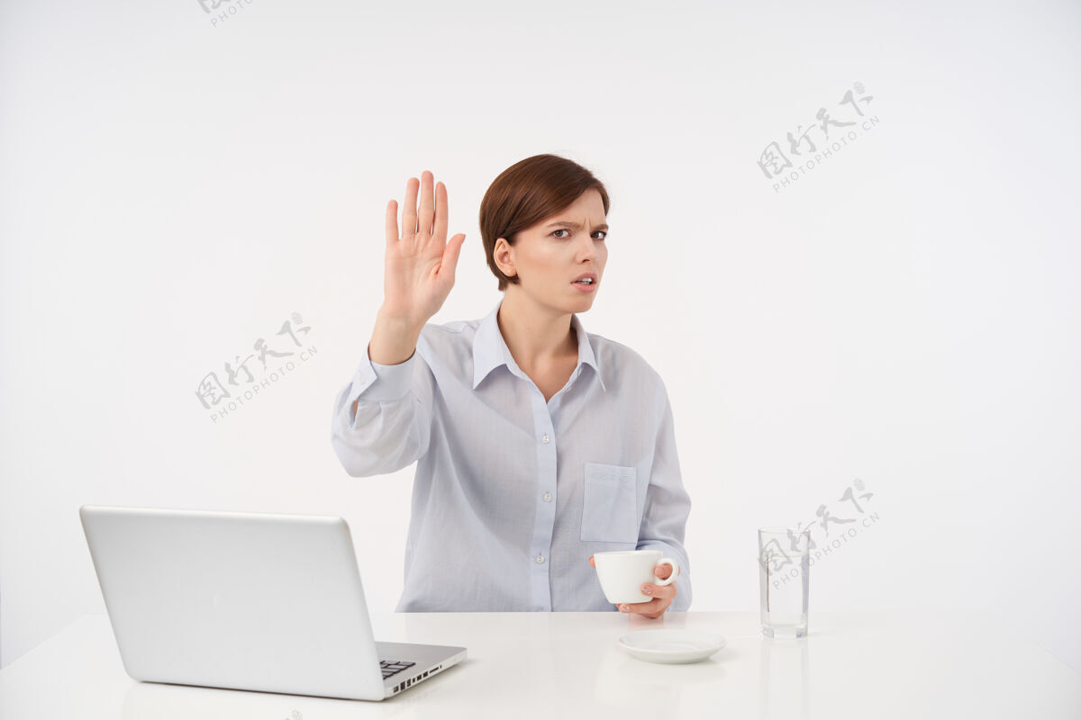员工室内画像：棕色头发的年轻女子 随意的发型 举着陶瓷杯 举着手掌做着停止的姿势 眉毛皱着 脸上带着不高兴的表情 坐在白色的椅子上拒绝化妆品玻璃