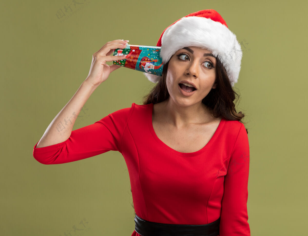 年轻好奇的年轻漂亮女孩戴着圣诞帽拿着圣诞咖啡杯在耳边听着谈话看着身边咖啡新年快乐