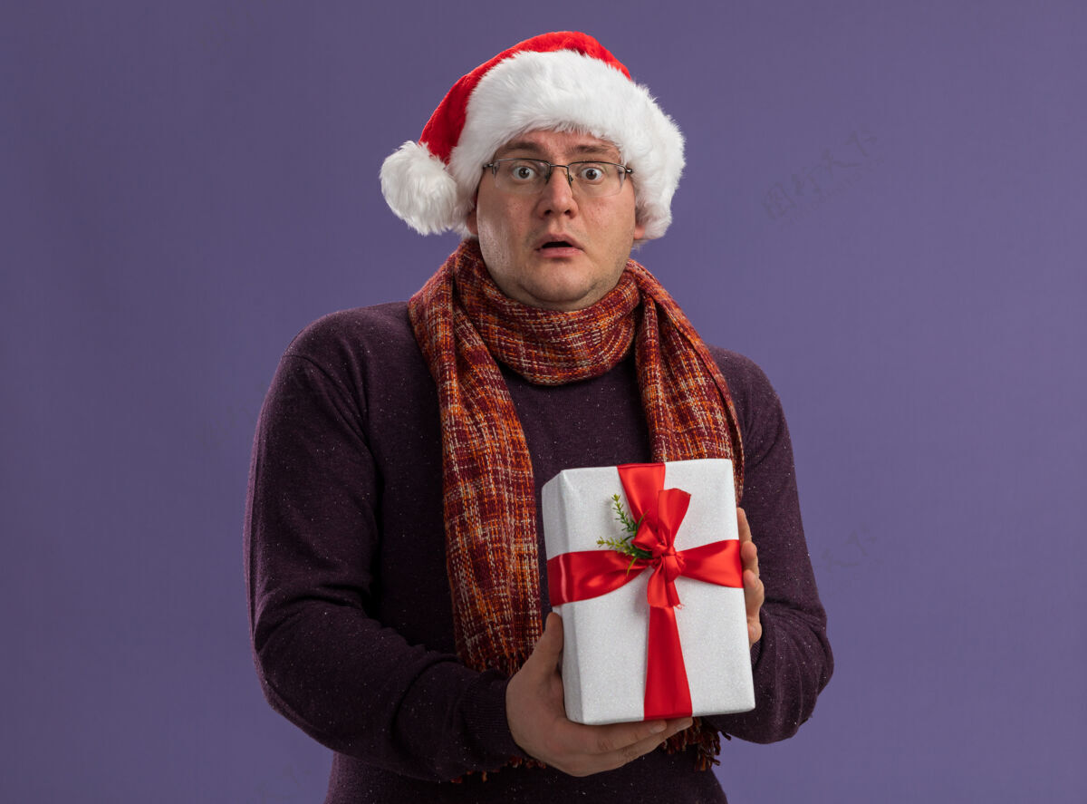 脖子给人印象深刻的成年男子戴着眼镜和圣诞帽 脖子上围着围巾 手里拿着礼包 看着隔离在紫色背景上的相机帽子快乐成人