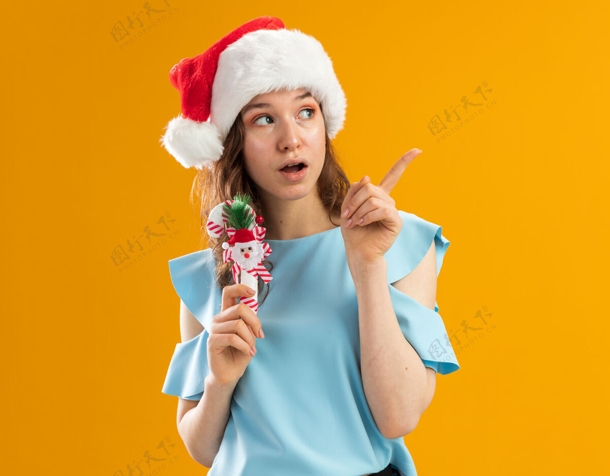 圣诞快乐身穿蓝色上衣 戴圣诞帽的年轻女子手持圣诞糖果手杖 惊讶地用食指指着什么东西新年帽子年轻