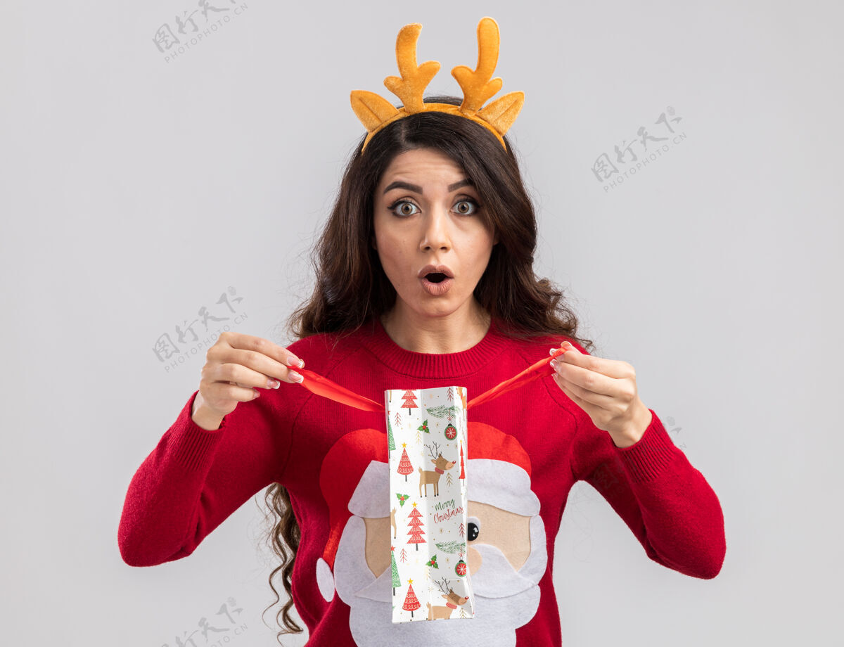 新年好奇的年轻漂亮女孩戴着驯鹿鹿角头带和圣诞老人毛衣拿着圣诞礼物包打开看圣诞老人礼物戴