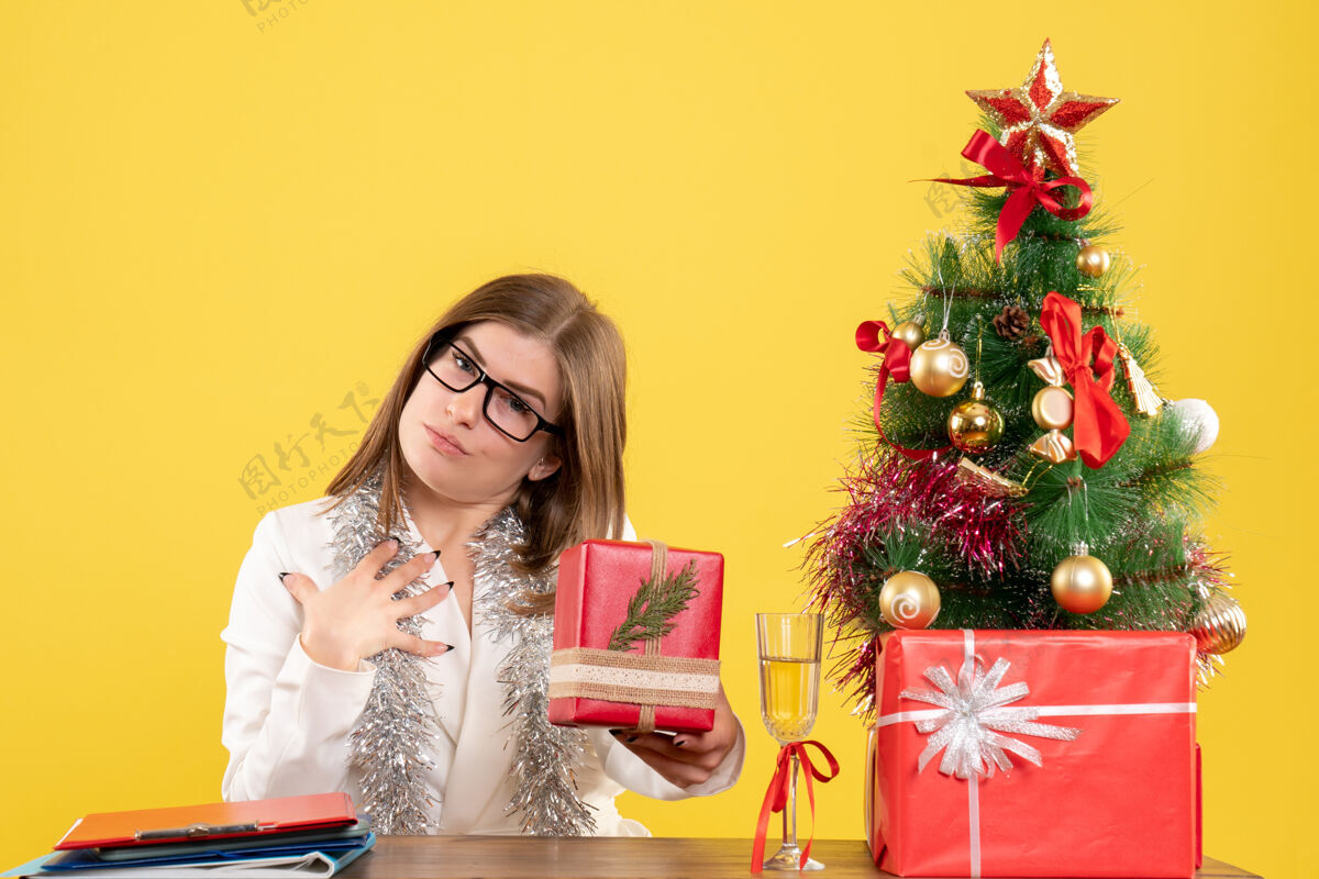 快乐正面图：女医生坐在桌子前 拿着礼物 黄色背景上的圣诞树和礼盒医生成人女医生