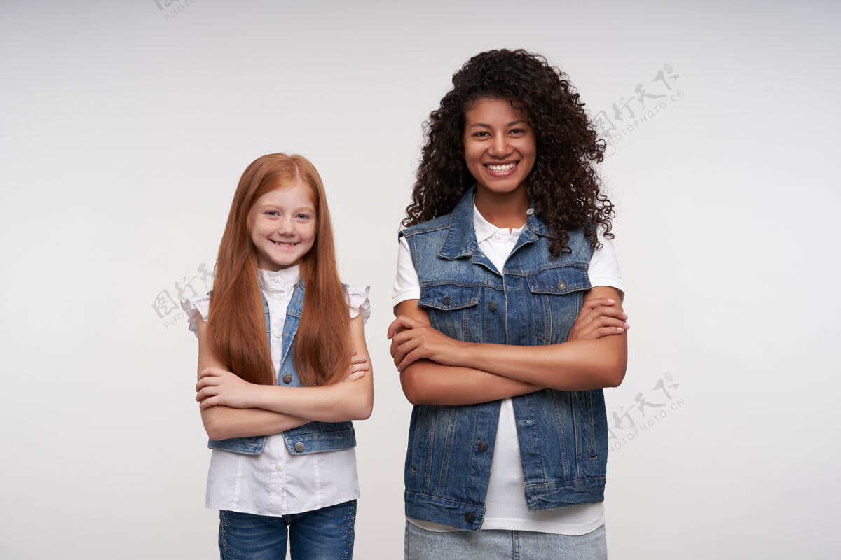 女人快乐的年轻黑皮肤黑发女性和红发长发漂亮女孩交叉双手的肖像 站在白色的椅子上开心地微笑着年轻手站立