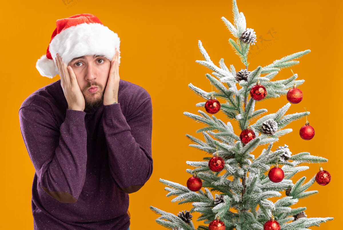 站着年轻人穿着紫色毛衣 戴着圣诞帽 看着相机迷茫而焦急的旁边 圣诞树的背景是橙色的帽子圣诞老人非常