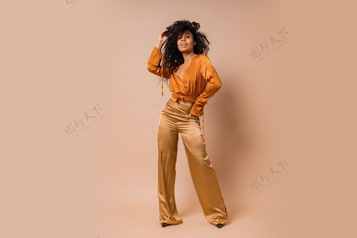 诱惑身着橙色上衣和金色丝绸长裤的优雅女士在米色墙壁上摆姿势高跟鞋令人惊叹的波浪形头发全长丝绸非洲肖像