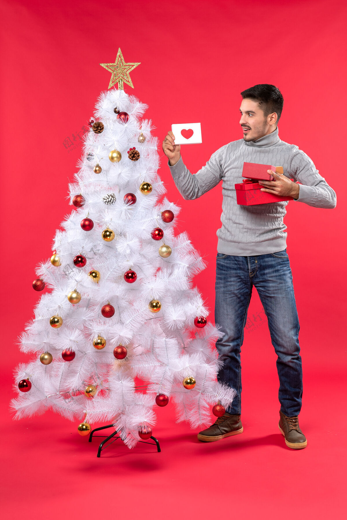 年轻人俯视图：穿着灰色上衣的年轻人站在装饰过的白色圣诞树旁 手里拿着红色的礼物衣服睡衣站立