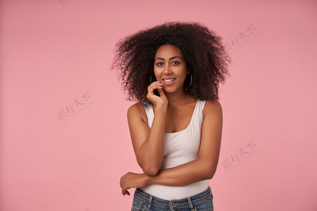 暗色快乐的年轻黑皮肤女性 随意的发型 抬起手轻轻抚摸着脸 带着迷人的微笑 穿着粉色的白衬衫和牛仔裤摆姿势发型积极面部