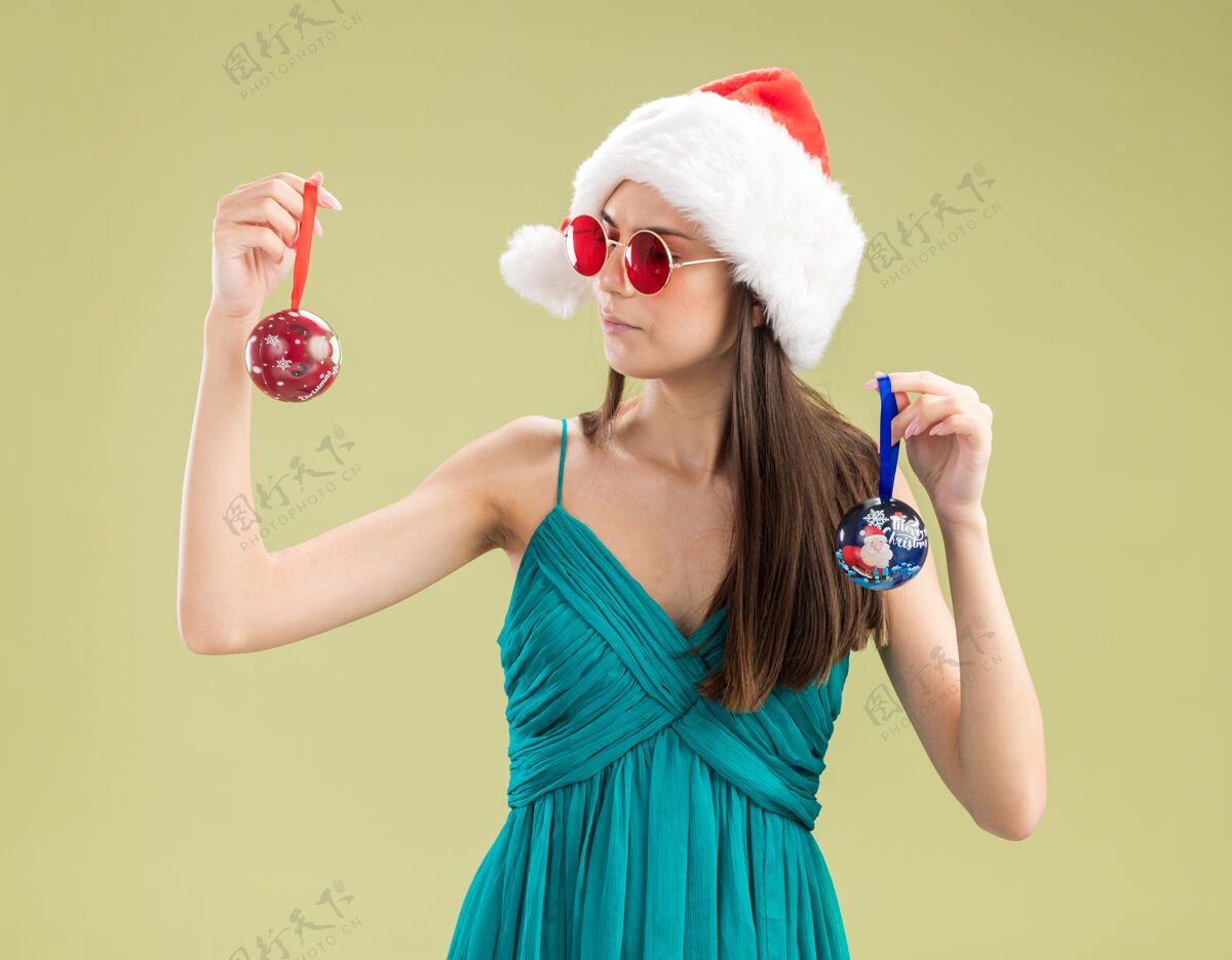 快乐自信的年轻白人女孩戴着太阳眼镜 戴着圣诞帽 看着玻璃球饰品圣诞老人新年女孩