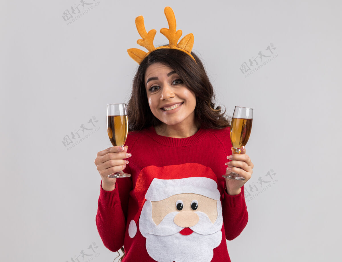 圣诞老人微笑的年轻漂亮女孩戴着驯鹿鹿角头带和圣诞老人毛衣拿着两杯香槟看着香槟鹿角年
