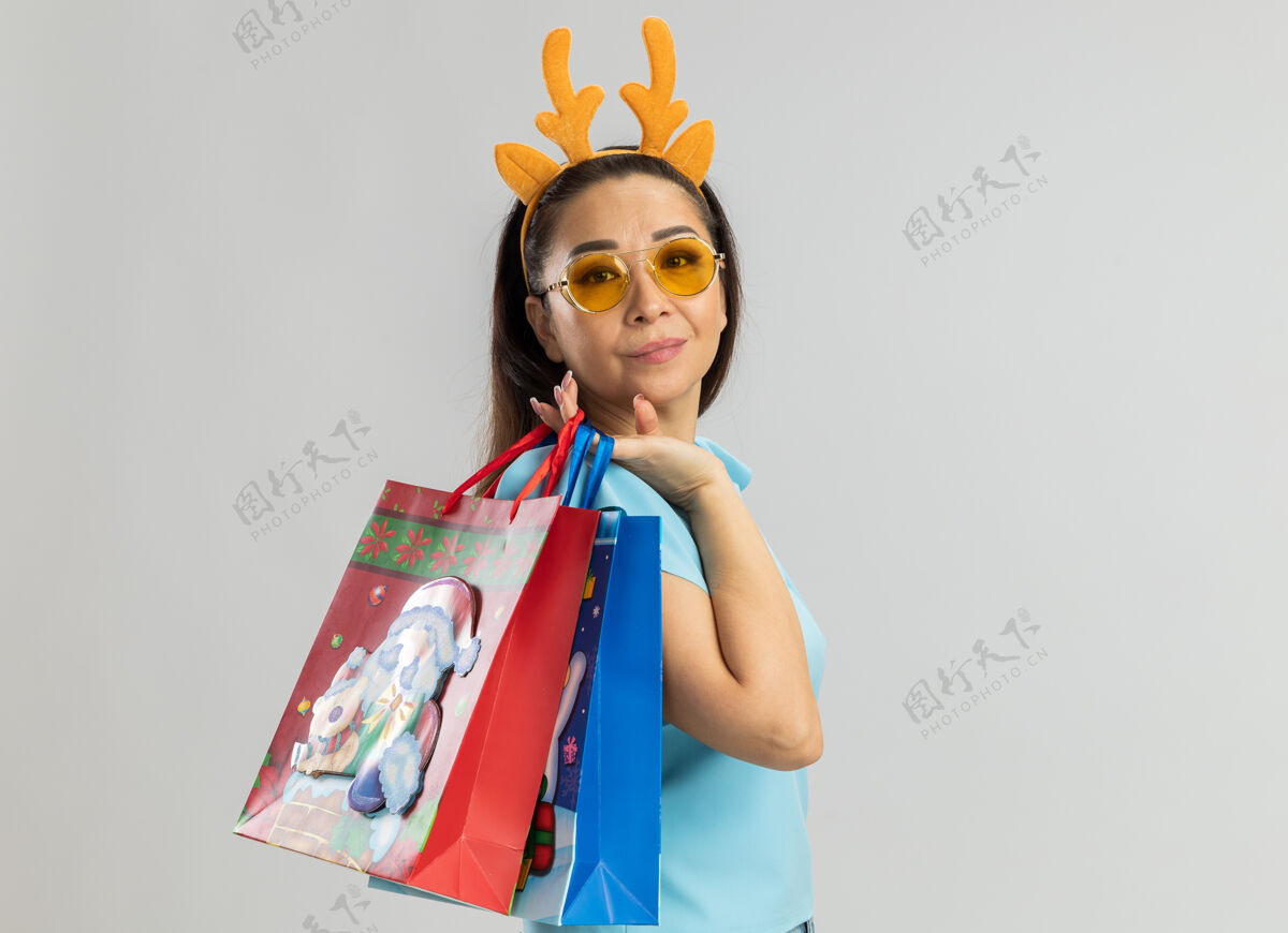 纸身穿蓝色上衣的年轻女子 戴着滑稽的鹿角边和黄色眼镜 手里拿着装有圣诞礼物的纸袋 看上去笑容满面 自信满满女人圣诞快乐年轻