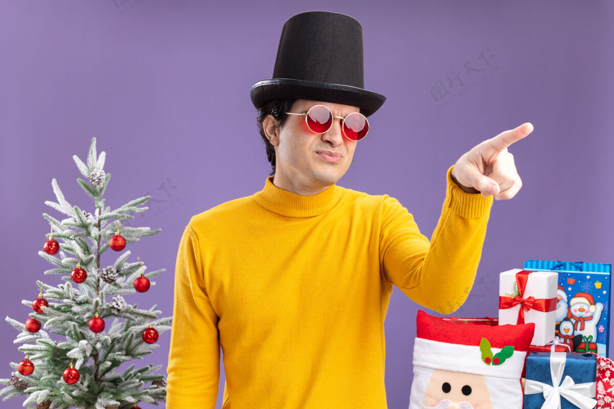 看一个穿着黄色高领毛衣 戴着黑色帽子的年轻人 不高兴地用食指指着站在圣诞树旁边的东西 紫色背景上的礼物圣诞节不高兴食指
