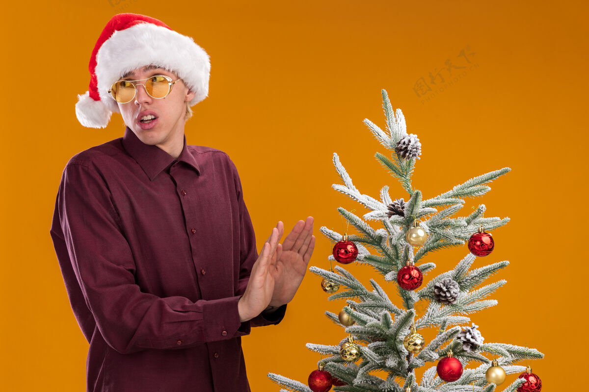 做一个戴着圣诞帽和眼镜的年轻金发男人站在装饰好的圣诞树旁 在橙色的背景下看着一边做着拒绝的手势男人未发布年轻人
