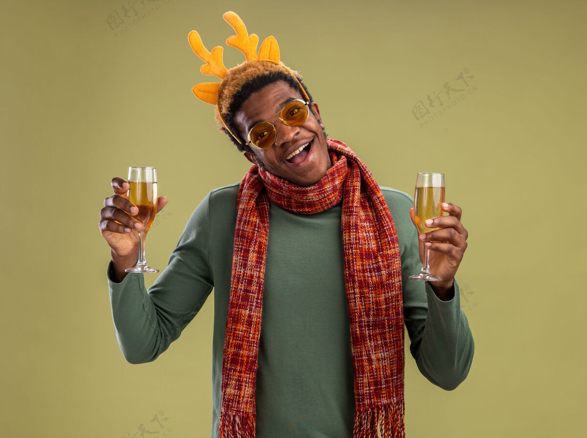 男人幸福的非洲裔美国人 戴着滑稽的边 戴着鹿角 脖子上围着围巾 手里拿着两杯香槟 站在绿色的墙上 开心地笑着围巾边缘鹿