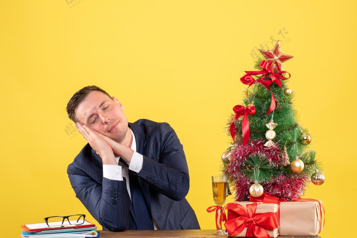 桌子睡意朦胧的男人坐在圣诞树旁的桌子前 黄色的礼物行政人员职业黄色
