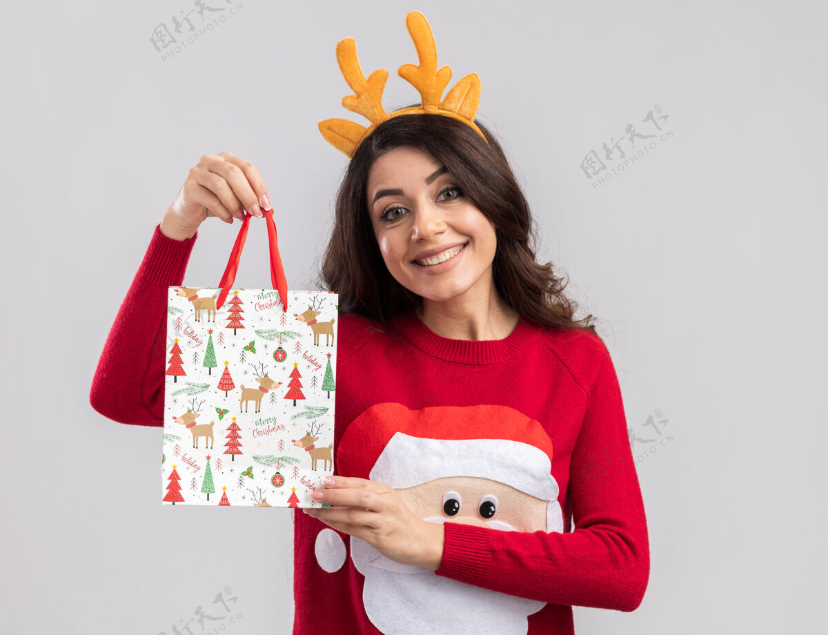 礼物微笑的年轻漂亮女孩戴着驯鹿鹿角头带和圣诞老人毛衣拿着圣诞礼物包看年驯鹿圣诞