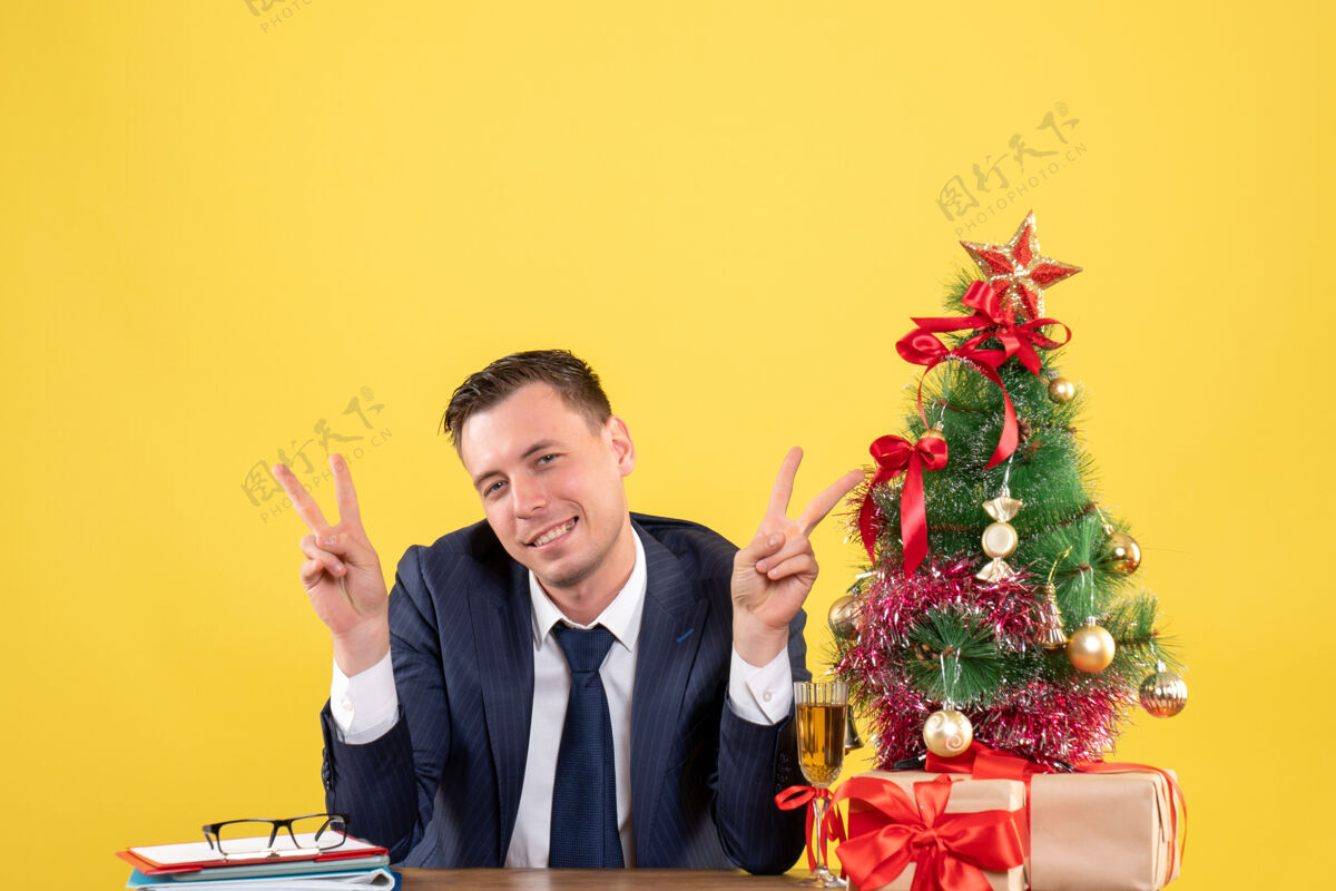 树圣诞树和黄色礼物旁的桌子旁 微笑着做人的胜利标志的正视图西装圣诞节行政人员