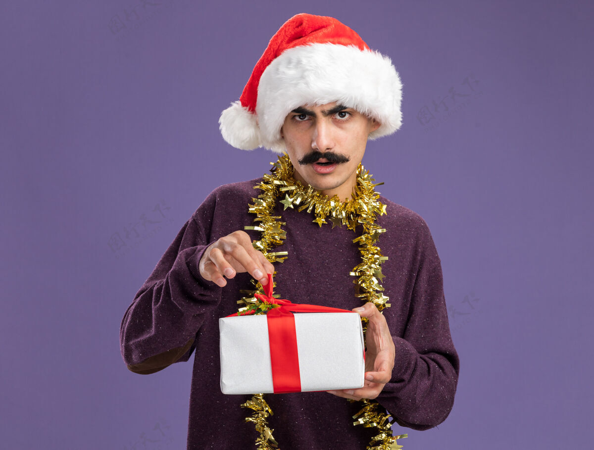 礼物一个留着胡子的年轻人戴着圣诞老人帽子 脖子上戴着金属丝 手里拿着圣诞礼物 严肃的脸站在紫色的背景上看着摄像机金属片快乐圣诞