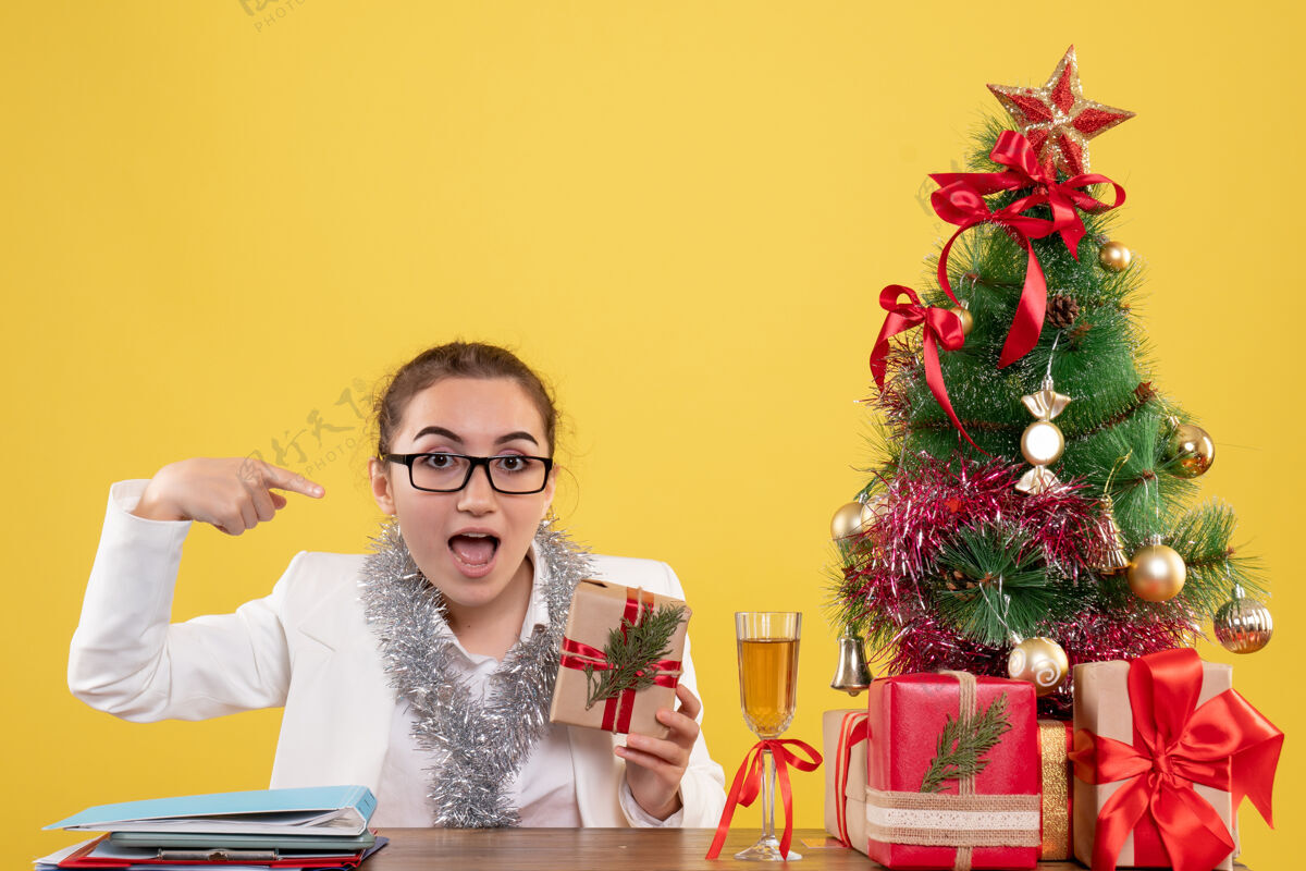 微笑前视图：女医生拿着圣诞礼物和黄色背景上的圣诞树坐着圣诞节背景礼物