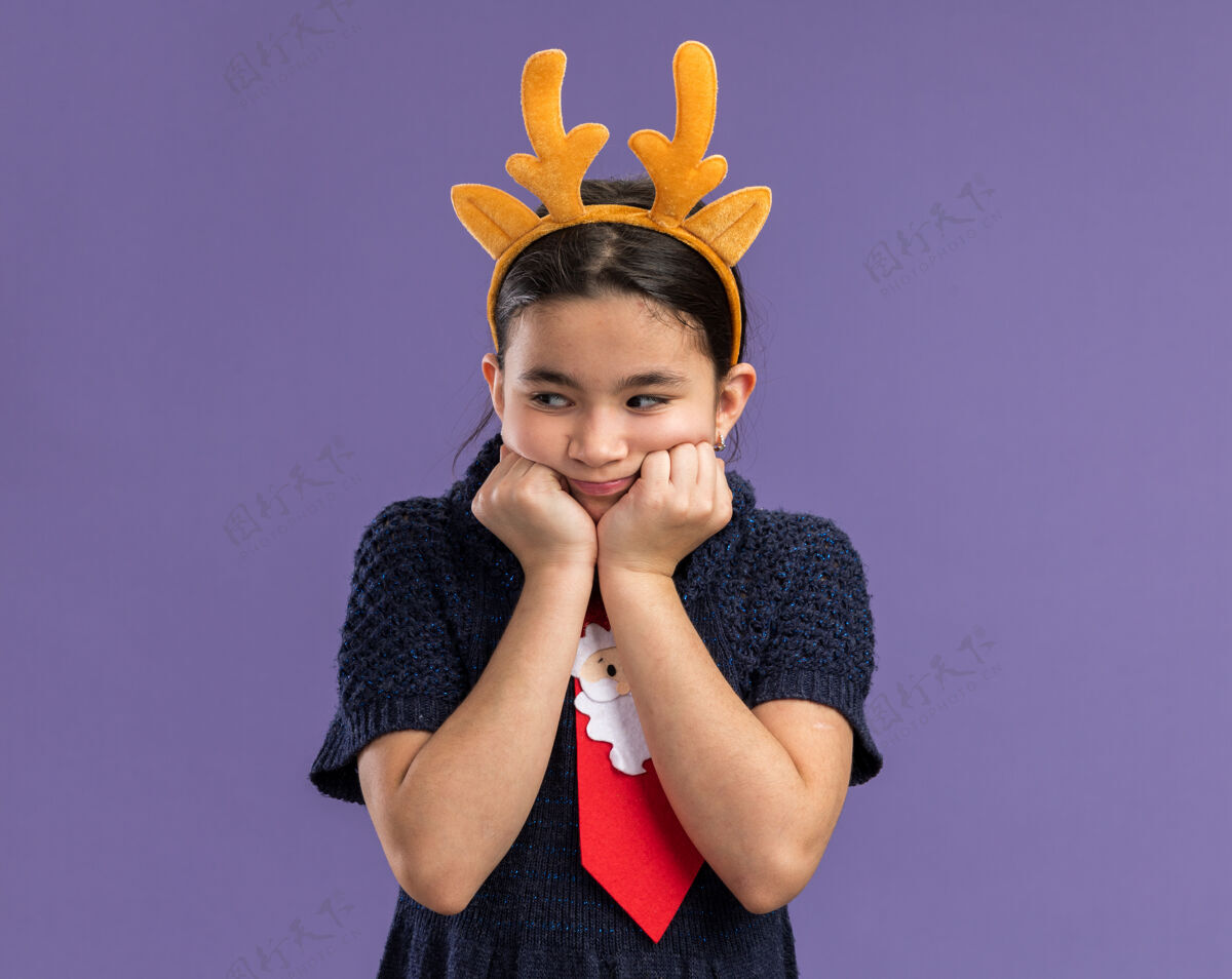 有趣穿着针织连衣裙的小女孩戴着红领带 边上有滑稽的镶边 头上戴着鹿角 站在紫色的背景上忧心忡忡地看着一边圣诞快乐领带边缘
