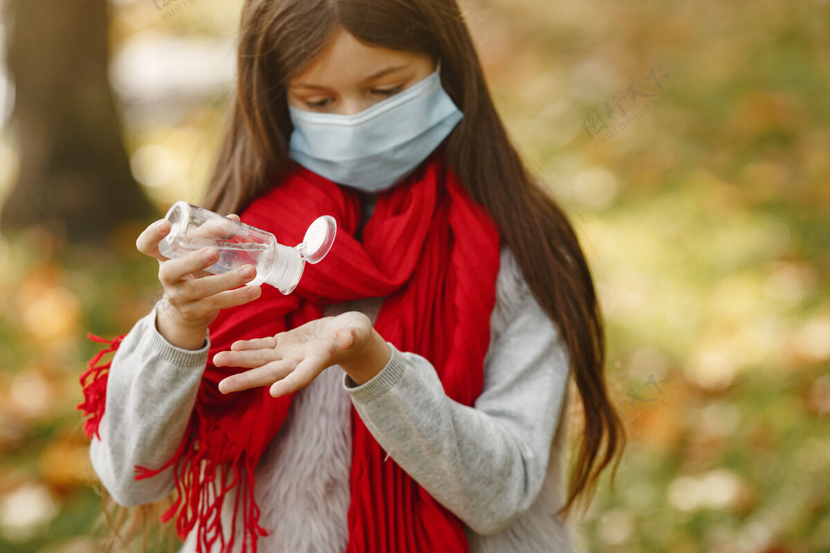 现代孩子站在秋天的公园里冠状病毒主题戴红领巾的女孩孩子用消毒棒安全围巾肖像