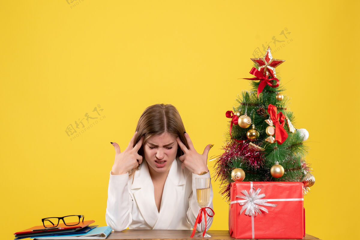 背景正面图：女医生坐在桌子前 在黄色背景下 戴着圣诞树和礼盒 感觉很累成人新女