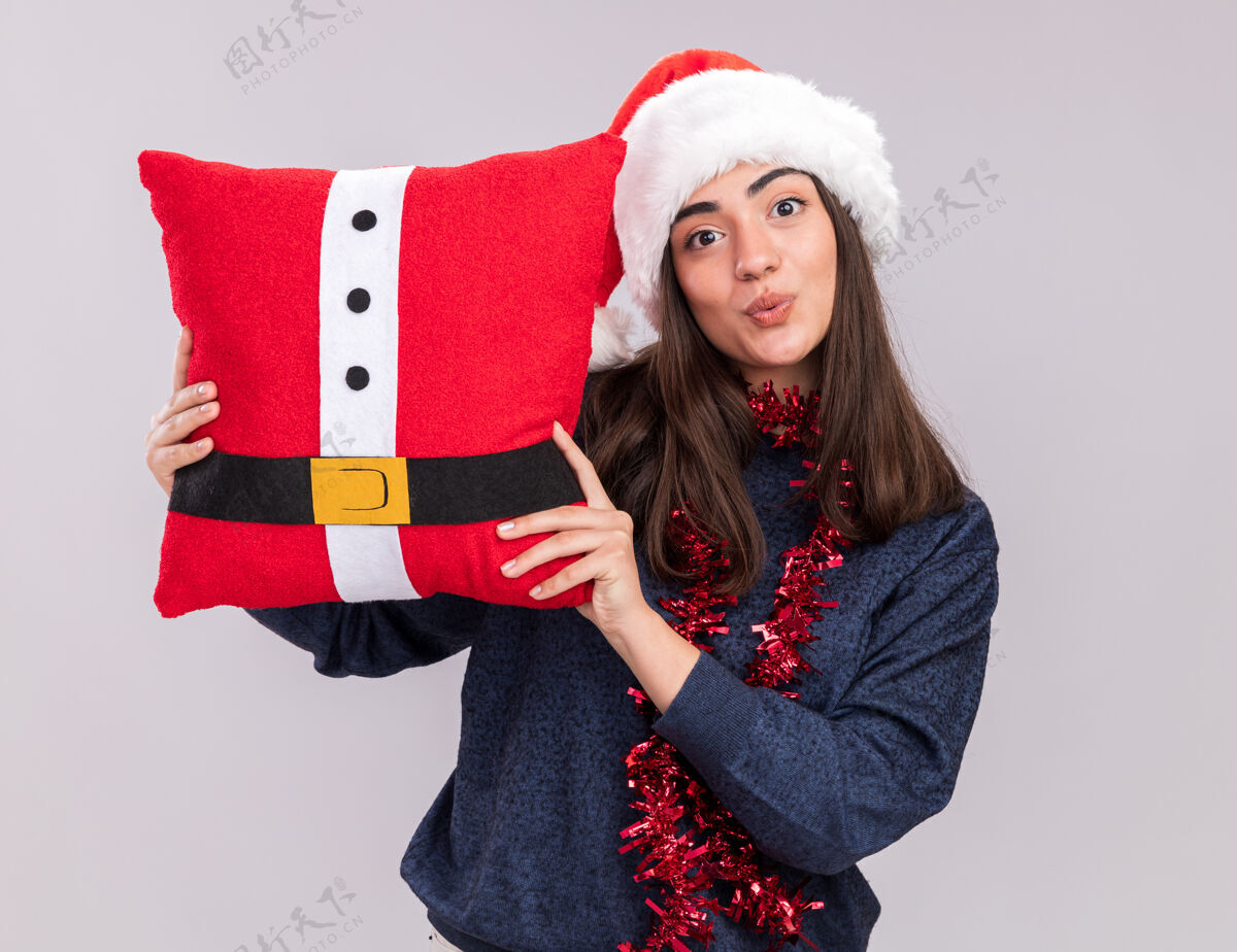 帽子令人印象深刻的年轻白人女孩戴着圣诞帽 脖子上戴着花环 拿着白色背景上的装饰枕头 并留有复制空间脖子圣诞老人年轻