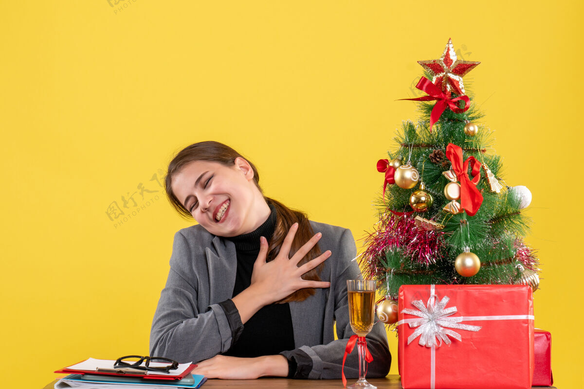 鸡尾酒前视图微笑的女孩坐在桌子旁 手放在胸前圣诞树和礼物鸡尾酒成人家手