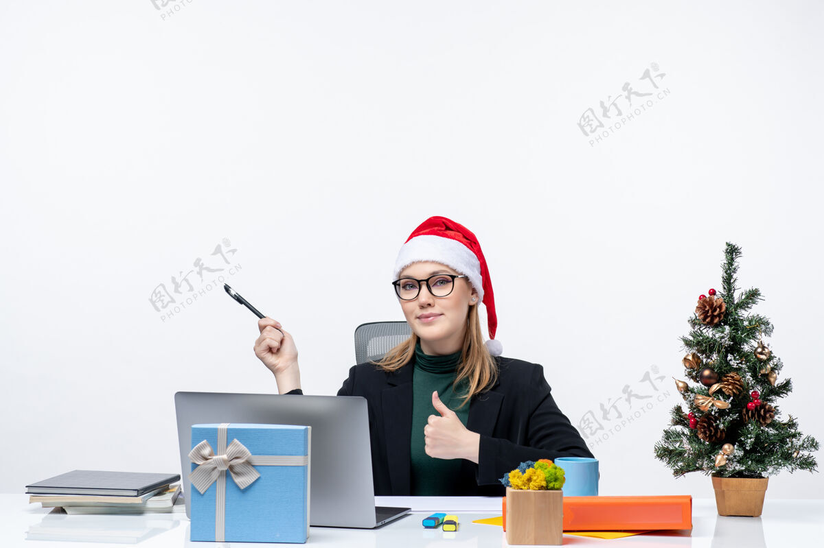 人戴着圣诞老人帽的金发女人坐在桌子旁 手里拿着圣诞树和礼物制作工作手势