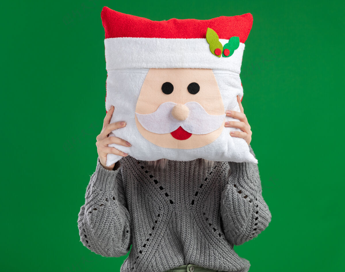 抱着穿着冬装毛衣的金发女郎拿着圣诞枕头遮住脸 枕头站在绿色的背景上金发站着圣诞节