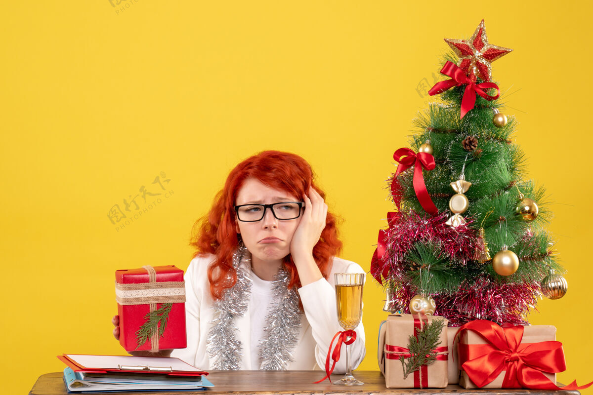 圣诞节前视图：女医生坐在桌子后面 黄色背景上有圣诞礼物花安排礼物