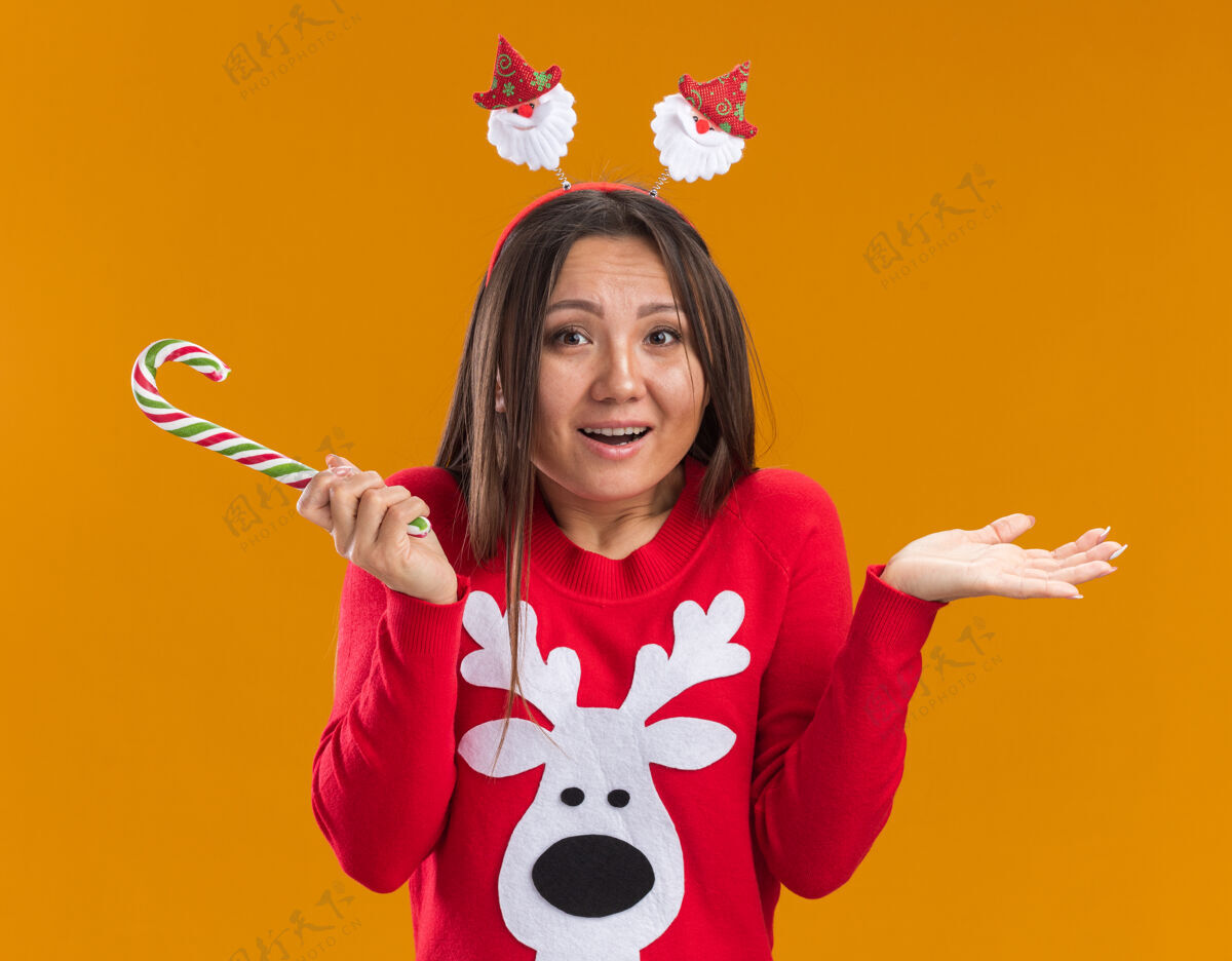 穿困惑的年轻亚洲女孩戴着圣诞发箍和毛衣拿着圣诞糖果摊开双手孤立在橙色的墙上蔓延年轻毛衣