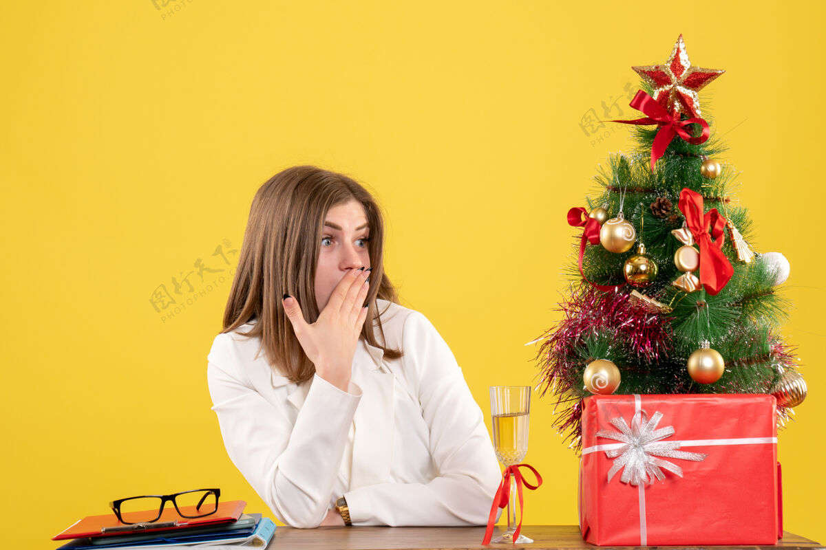 圣诞节正面图：女医生坐在黄色背景的桌子前 放着圣诞树和礼品盒背景职业成人