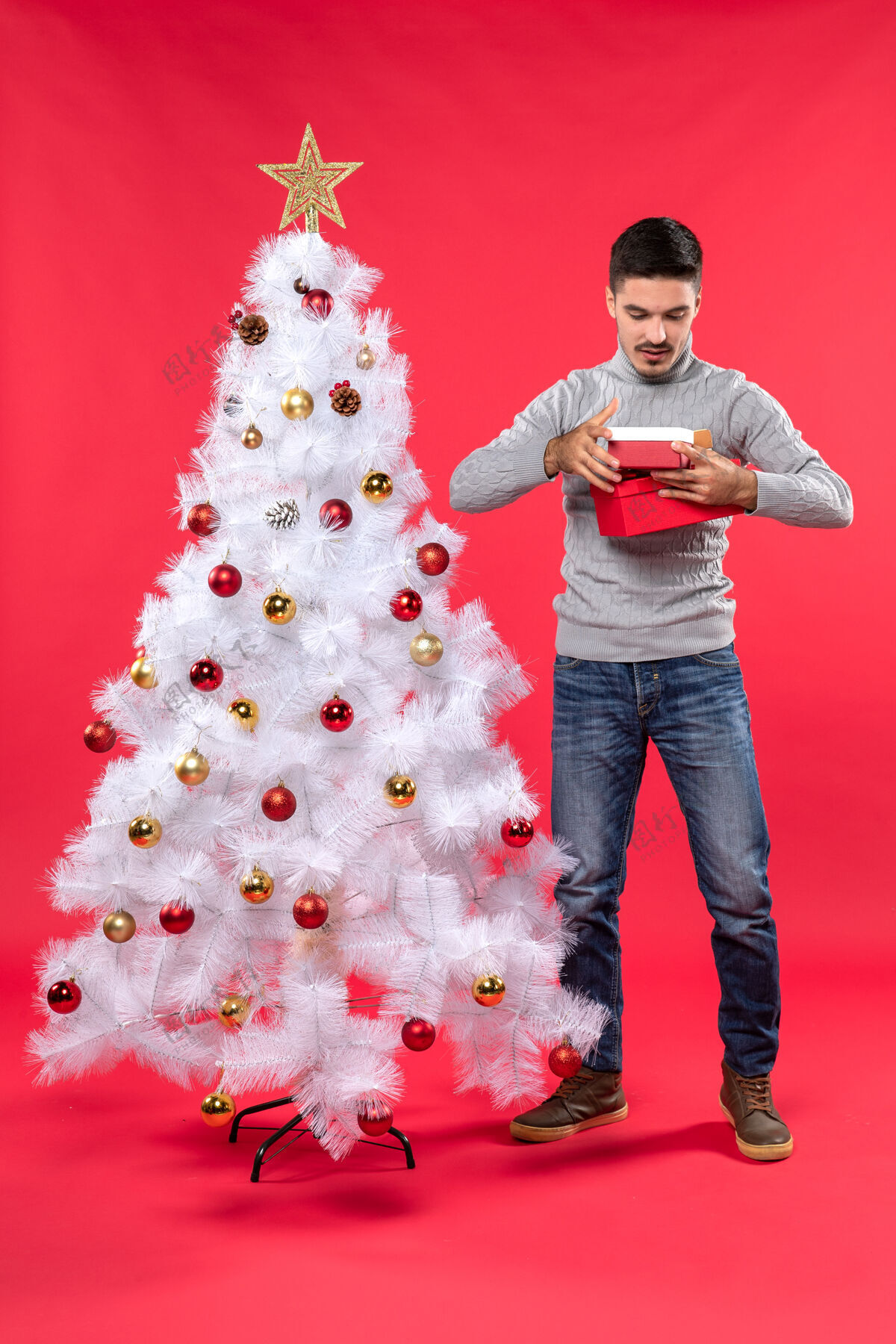 雪人一个穿着灰色上衣的浪漫成年人站在装饰过的白色圣诞树旁 手里拿着红色的礼物礼物衬衫欢快