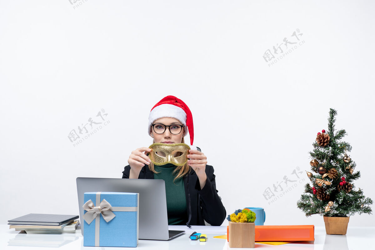 自由职业者戴着圣诞老人帽 眼镜和面具的年轻女子坐在桌子旁 手里拿着圣诞树和礼物礼物年轻人桌子