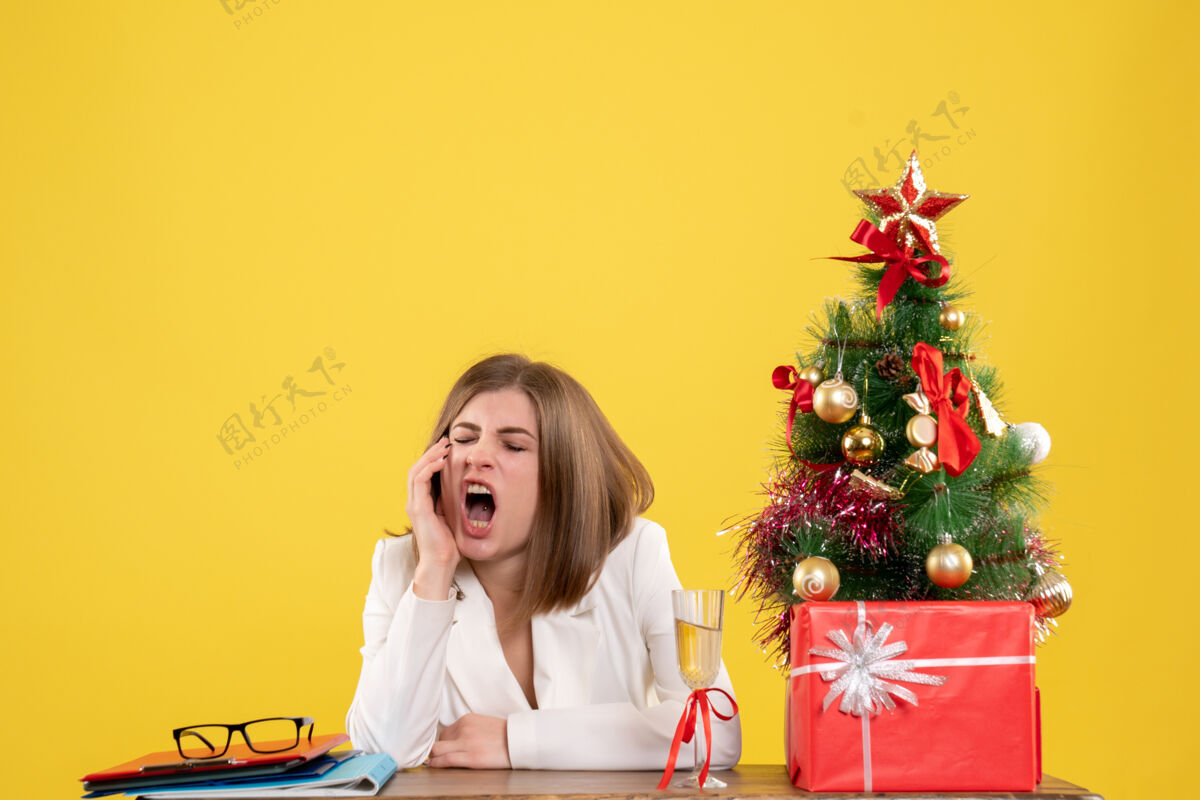 人正面图：女医生坐在桌子前打着哈欠 背景是黄色的圣诞树和礼盒快乐女性医院
