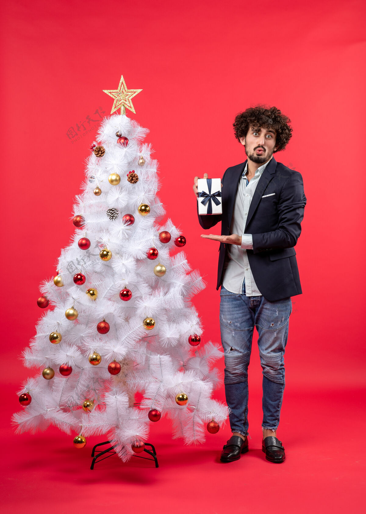 庆祝新年庆祝与震惊的年轻人展示他的礼物附近装饰白色圣诞树上的红色镜头男人震惊圣诞树